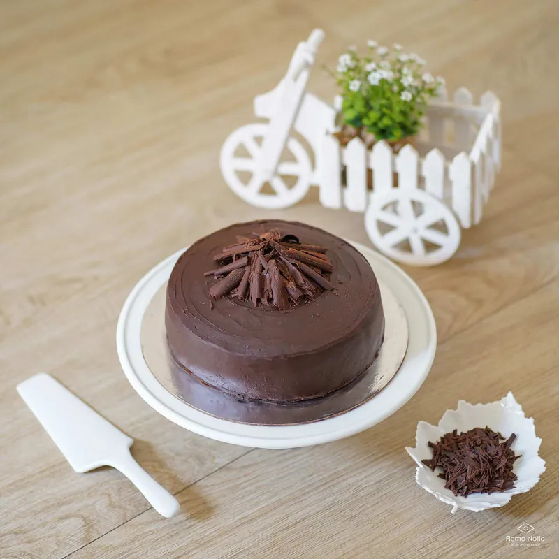 Chocolate cake with chocolate curls on a white cake stand, beside a little white bicycle planter and a cake server